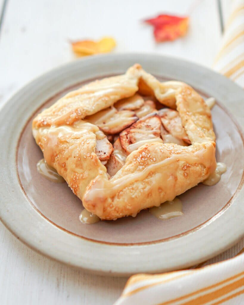 An individual apple galette with golden crust, apple filling, and a drizzle of caramel on a light brown plate
