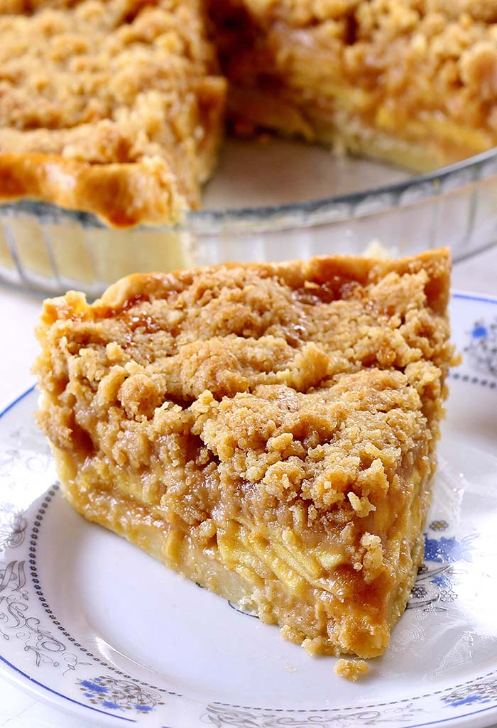 Slice of Dutch apple pie with a thick crumb topping on a decorative white and blue plate, with the rest of the pie visible…