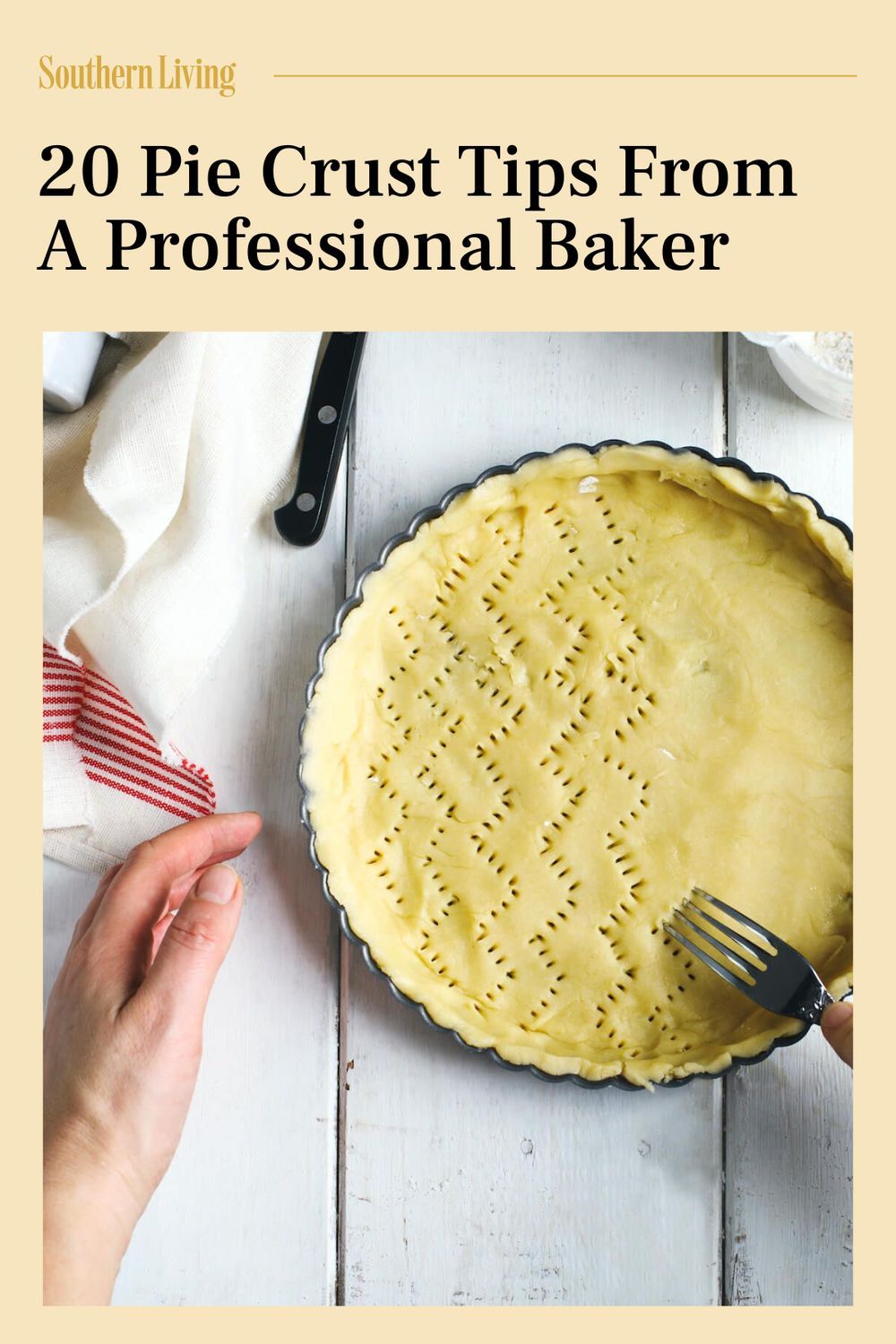 A hand uses a fork to prick holes into an unbaked pie crust in a metal pie pan, laid on a white wooden surface.