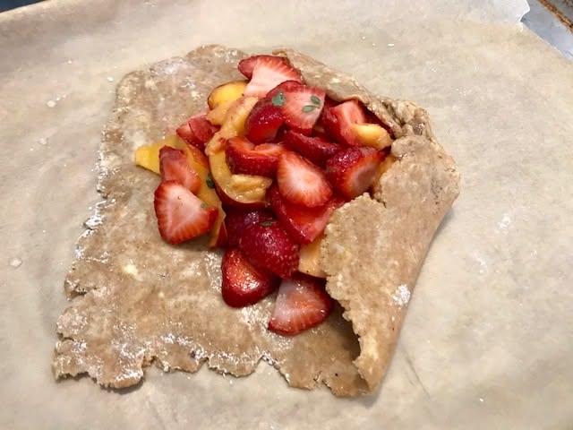 Unbaked galette on parchment paper, with sliced strawberries and peaches in the center, and the crust partially folded over…