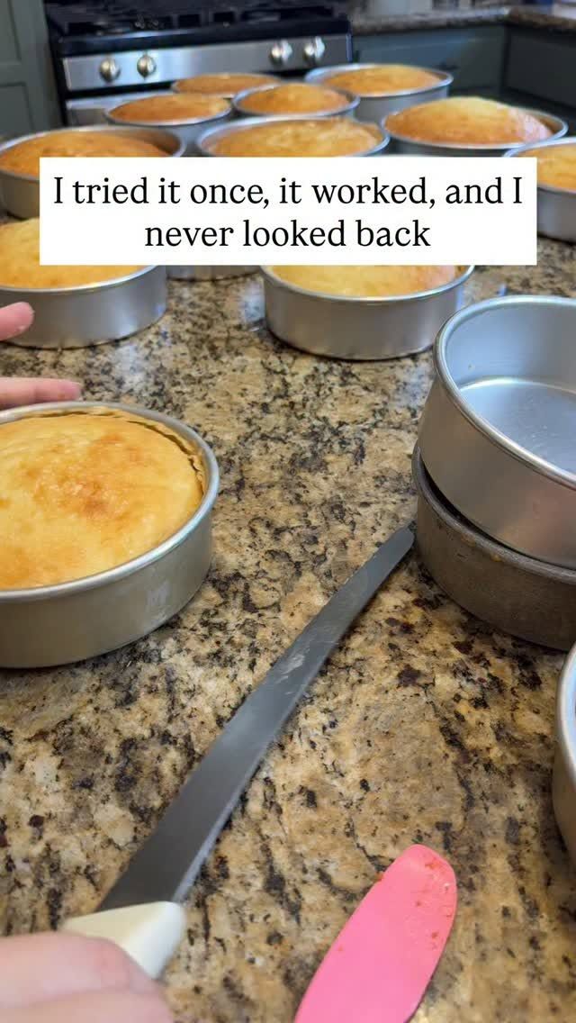 Several golden-brown cake layers in thin aluminum pans on a speckled kitchen counter next to a metal spatula and pink…