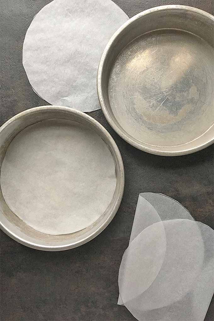 Two round metal cake pans, one lined with parchment paper. Several cut parchment circles are next to the pans.