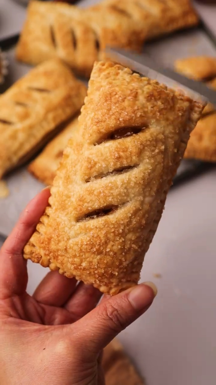 A hand holding a golden-brown apple turnover with crimped edges and sugar coating.