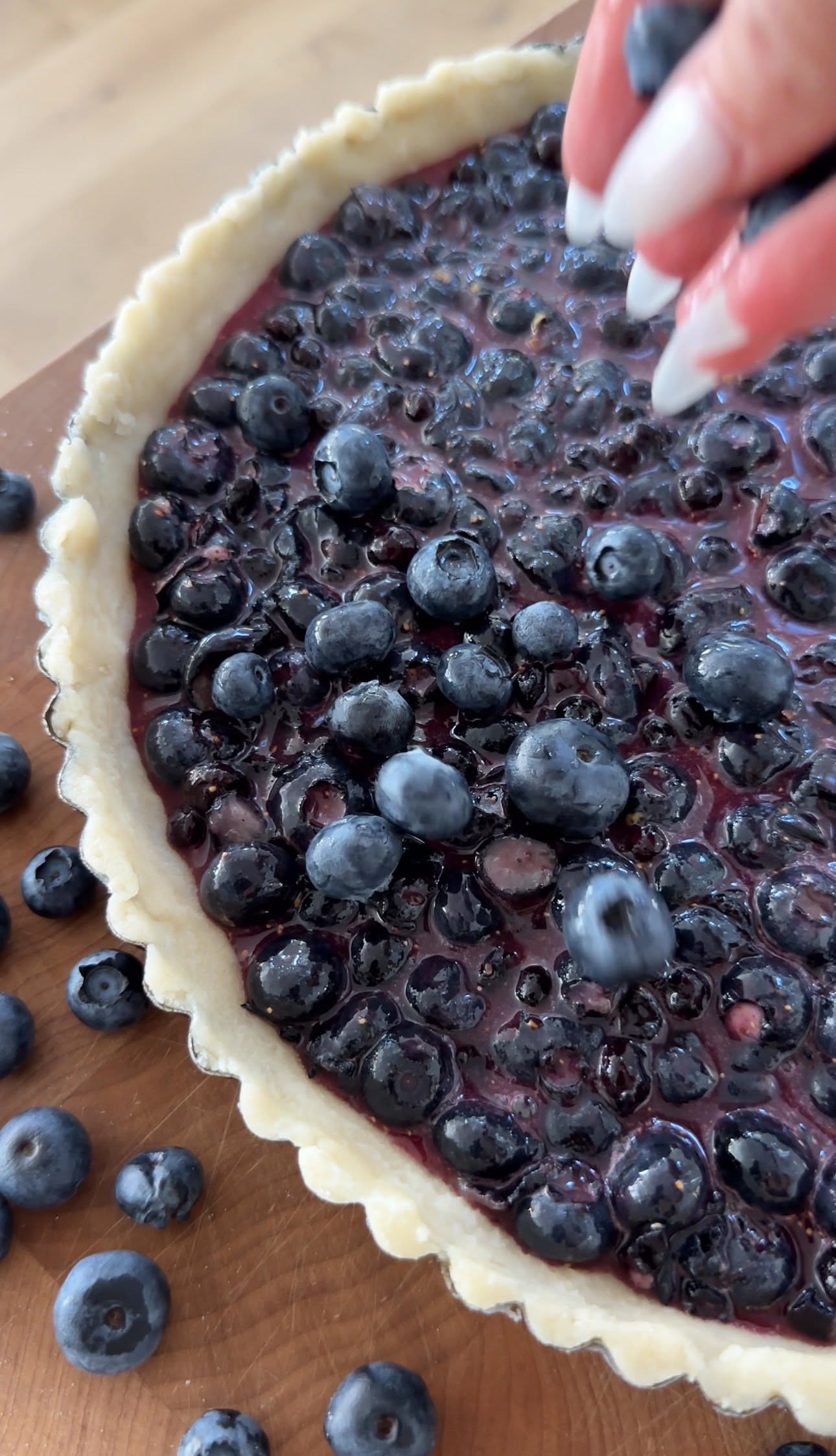 A hand placing fresh blueberries into a tart shell already filled with a deep purple blueberry mixture, with raw tart dough…