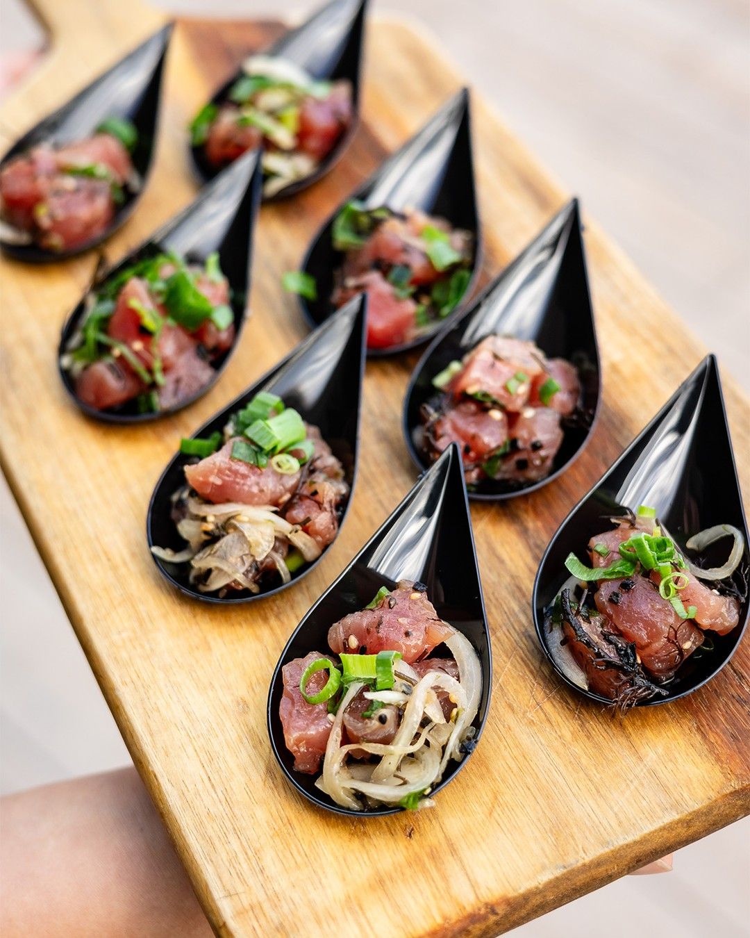 A wooden tray holds several black, tear-shaped spoons, each filled with diced tuna, sliced onions, and green herbs.
