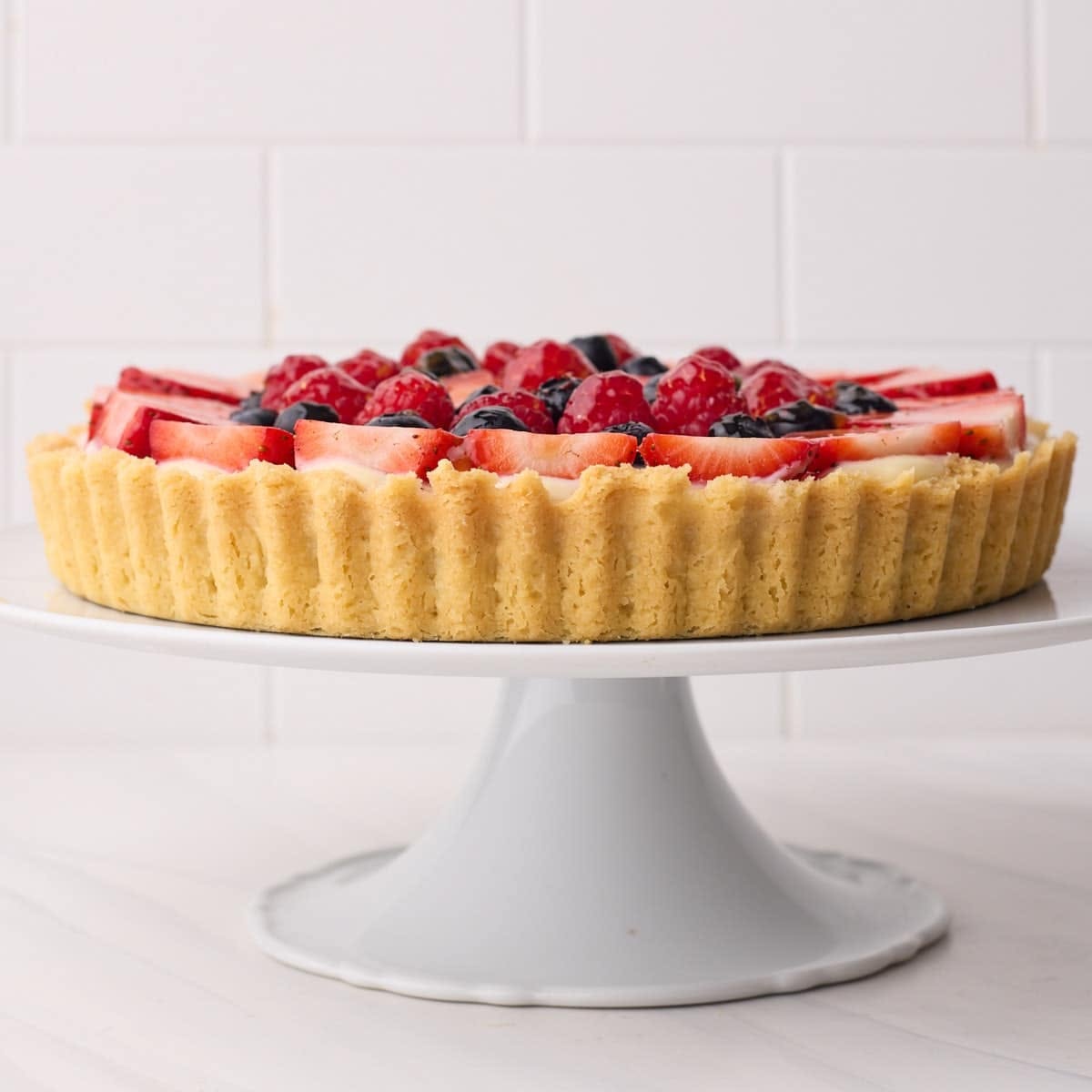 Close-up of a fruit tart with a golden, fluted crust filled with cream and fresh berries, on a white cake stand.