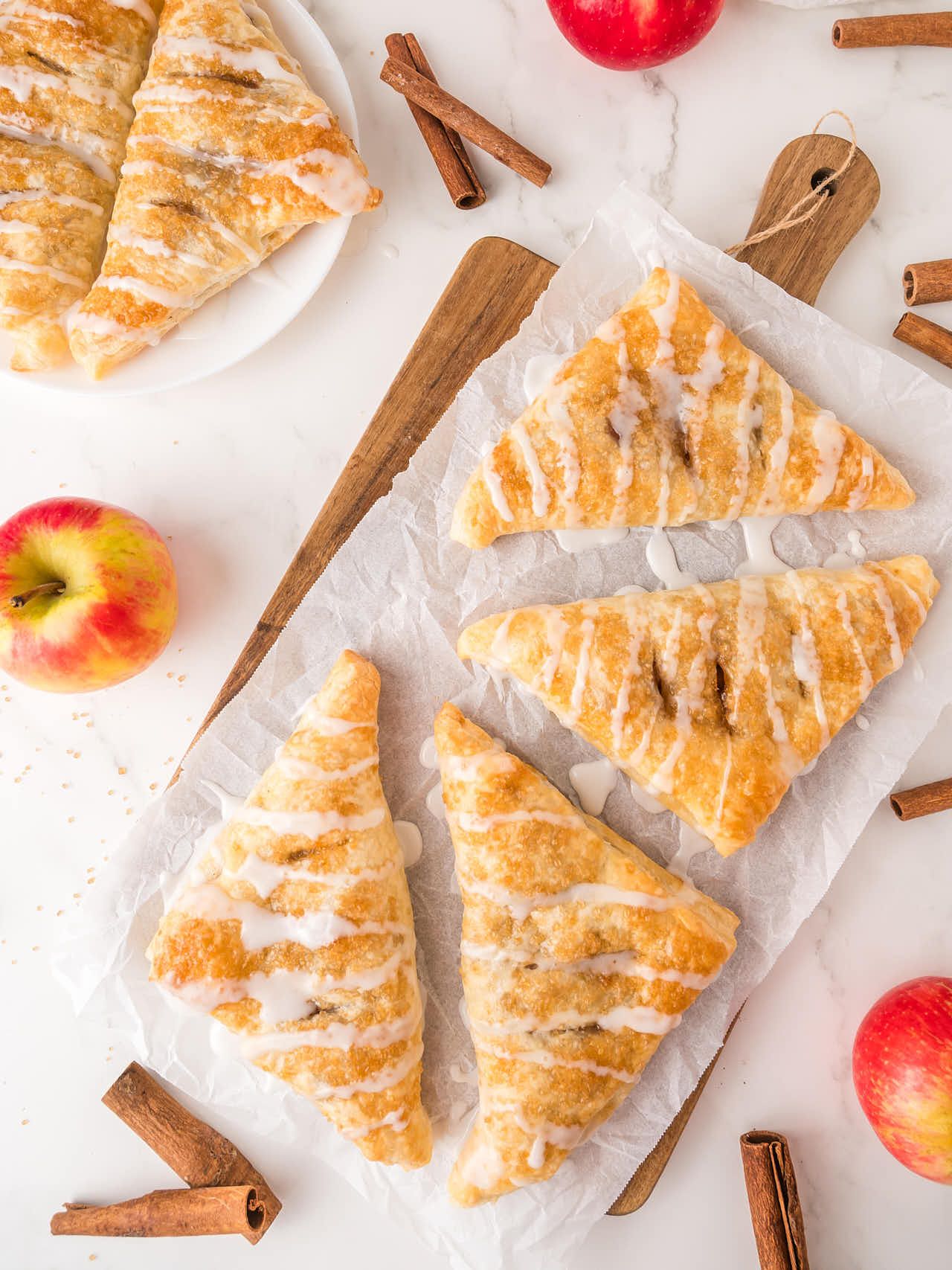 Several golden brown apple turnovers with white icing drizzle on parchment paper, next to whole apples and cinnamon sticks