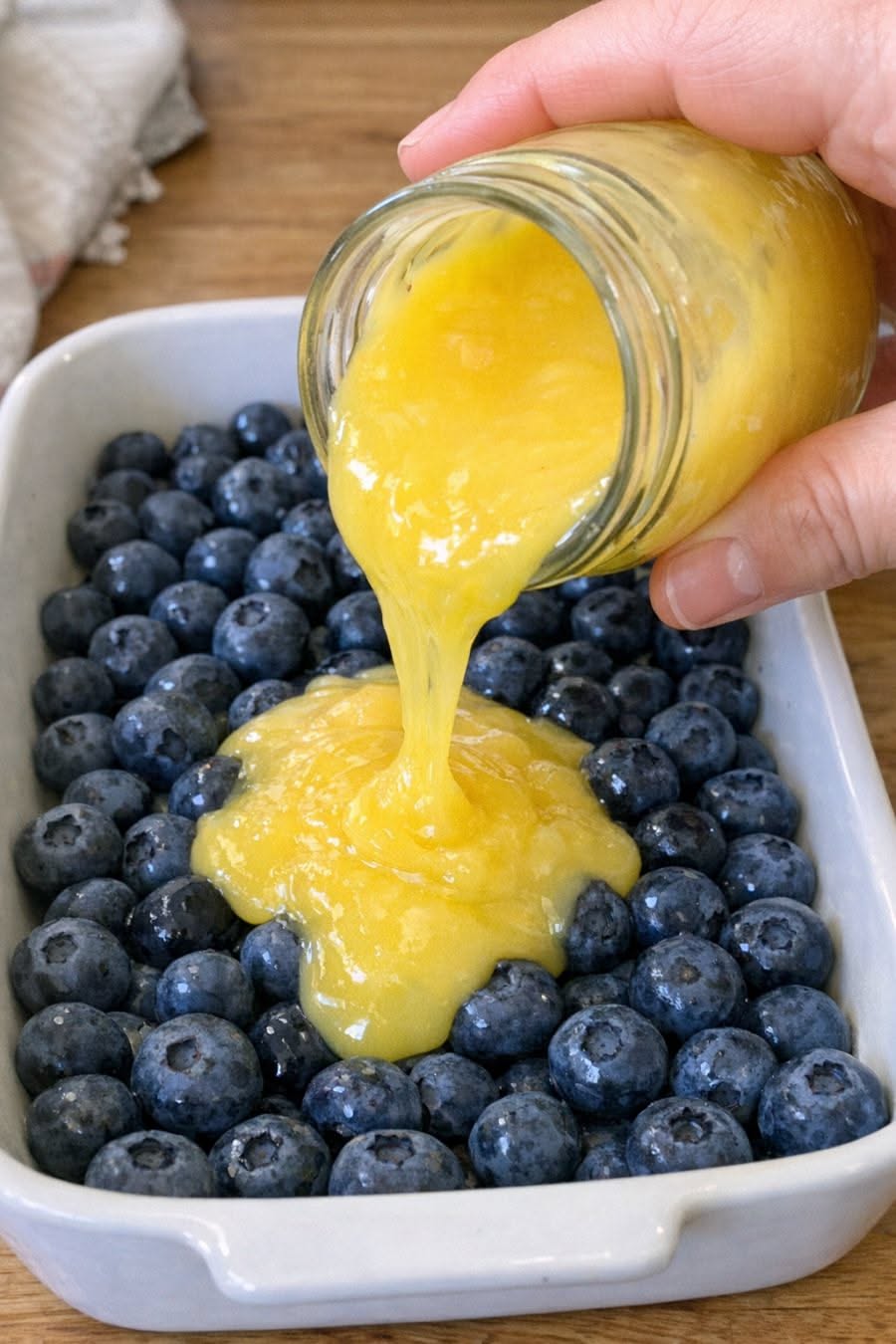 Lemon curd being poured from a glass jar onto a layer of blueberries in a white baking dish.
