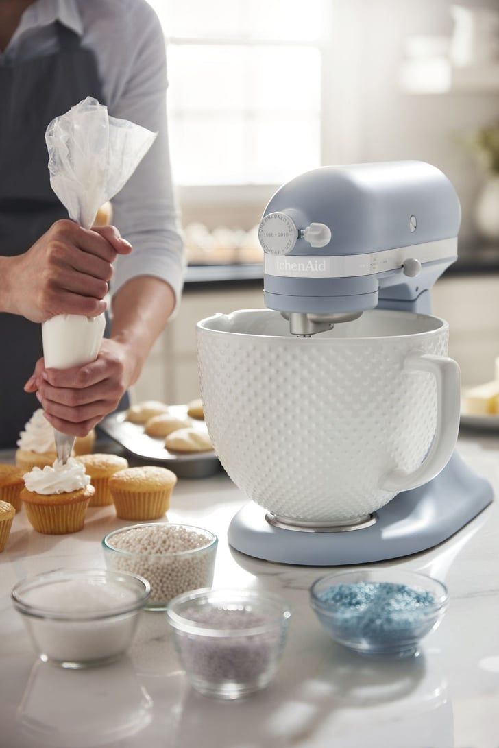 Person decorating a cupcake with frosting from a piping bag next to a light blue stand mixer with a textured white ceramic…