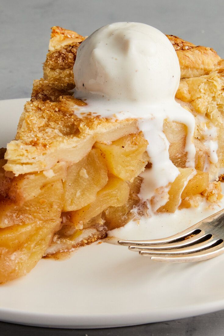 Close-up of a slice of apple pie topped with melting vanilla ice cream, beside a fork on a white plate.