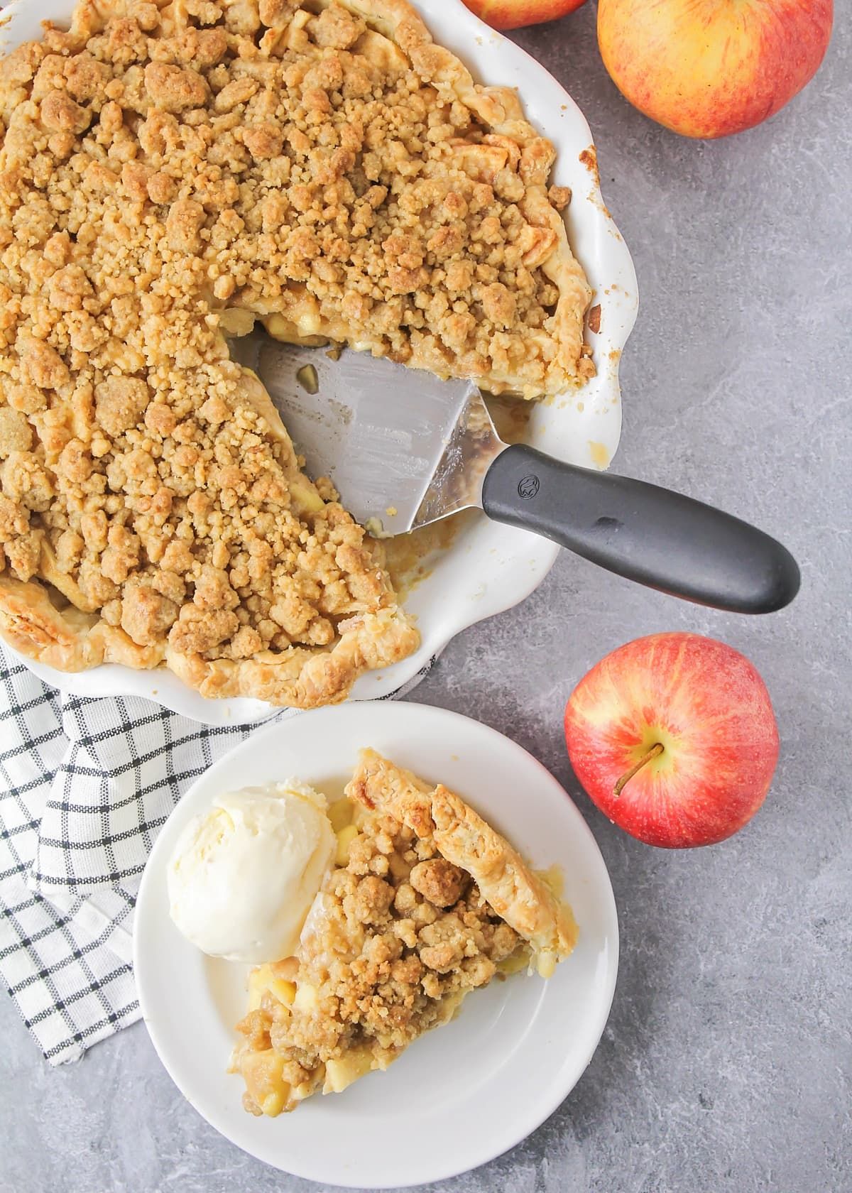 A slice of Dutch apple pie with ice cream on a white plate, next to a full pie in a dish with a server, and two apples.