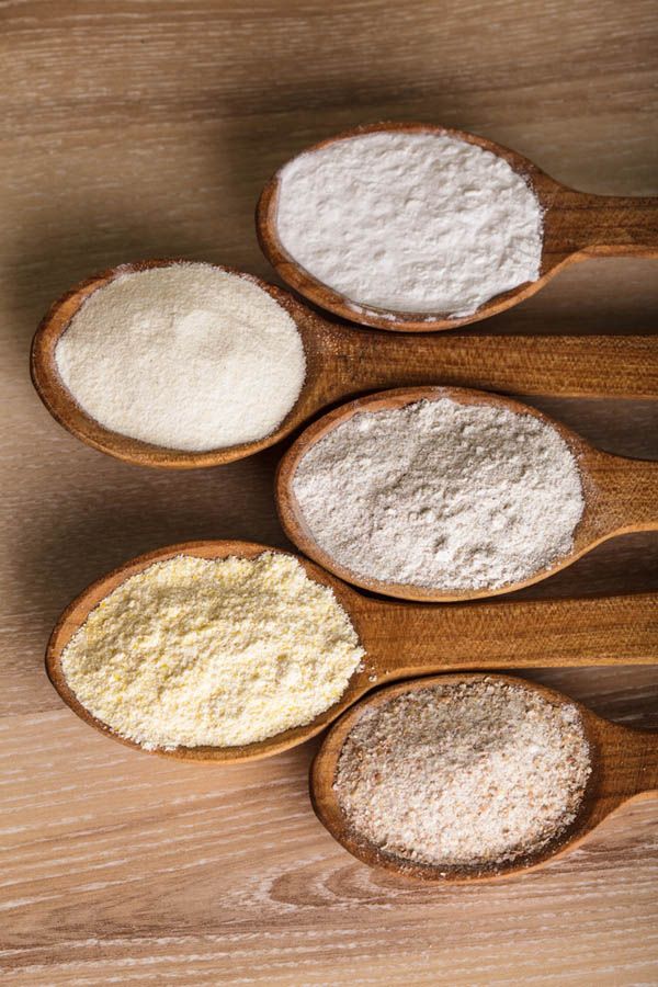 Five wooden spoons, each holding a different type of flour, arranged on a light wooden surface.