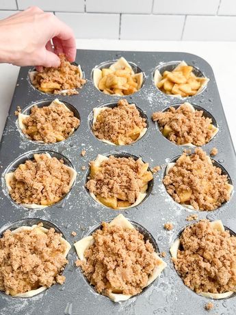 A hand sprinkles brown crumble topping over mini apple galettes in a muffin tin.