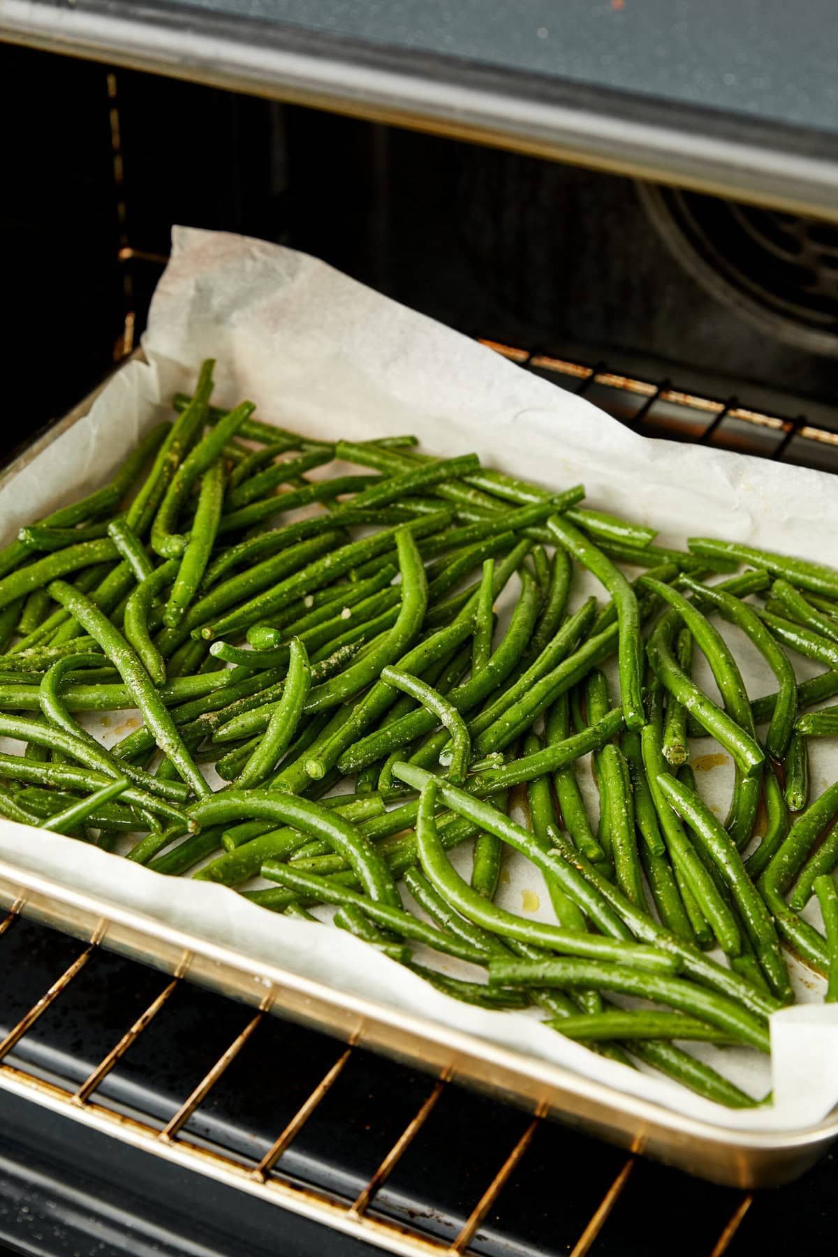 Green beans seasoned and spread on parchment paper on a baking sheet, inside an oven