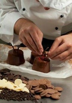 Chef's hands meticulously decorating individual chocolate desserts on parchment paper, with chocolate pieces and chips in…