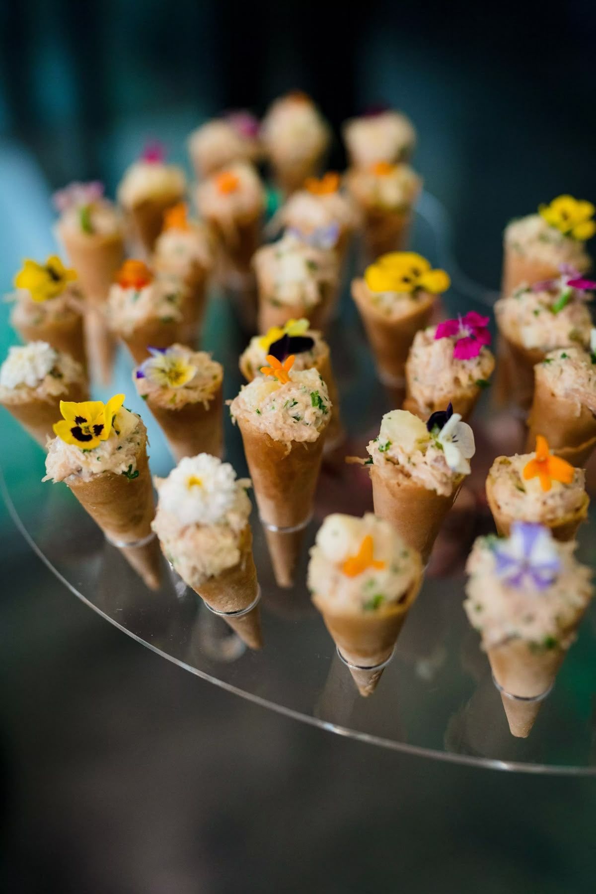 Several small savory cones, each filled with a creamy mixture and garnished with tiny edible flowers, arranged on a clear…
