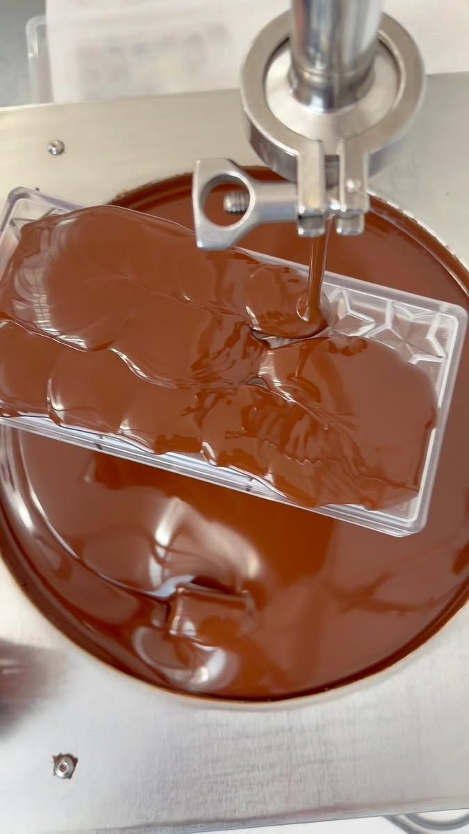 Melted chocolate pouring from a dispenser into a clear plastic mold, with a large vat of melted chocolate below.