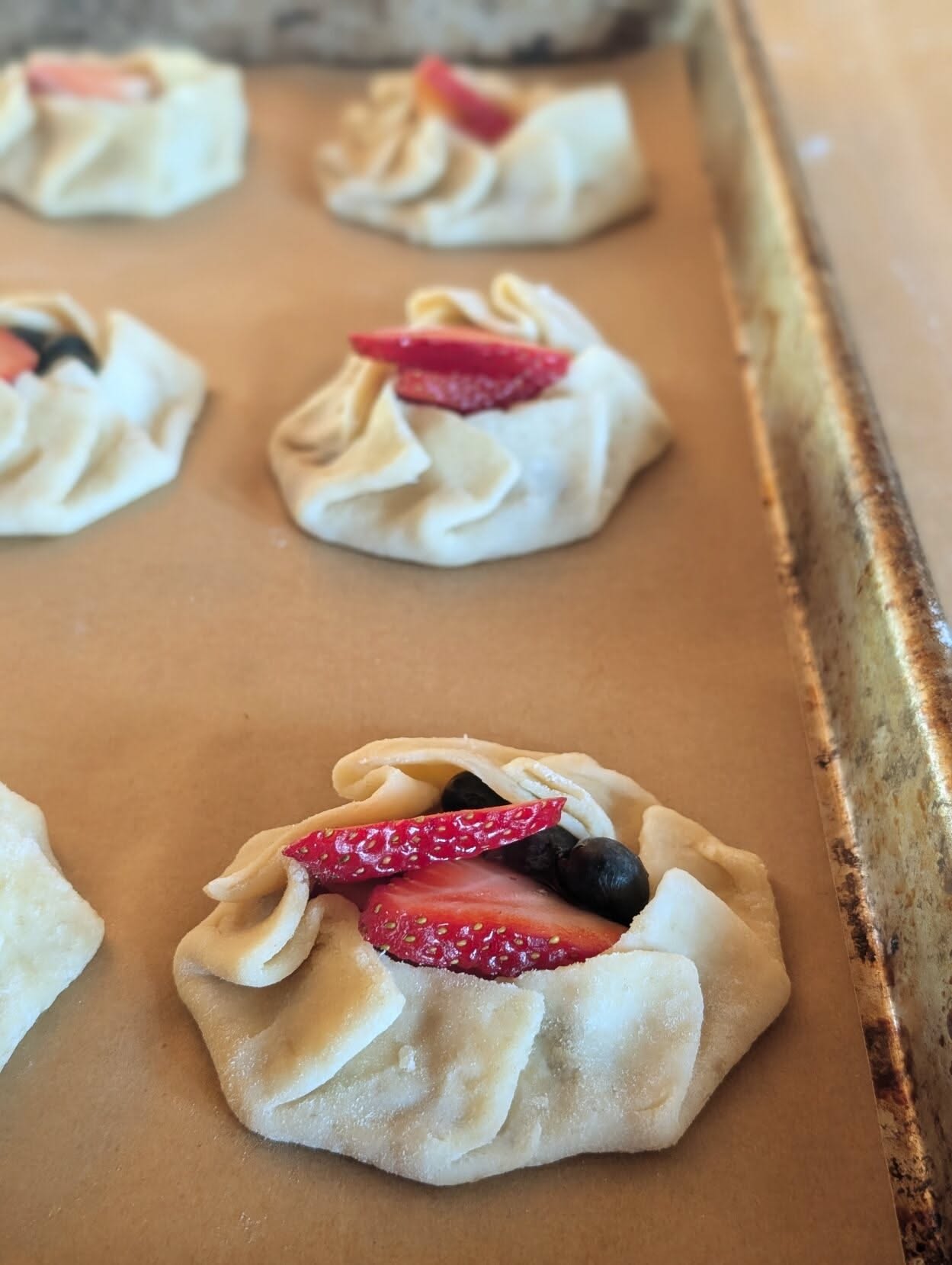 Close-up of several small, unbaked galettes on a baking sheet, with fruit filling visible and dough edges folded.