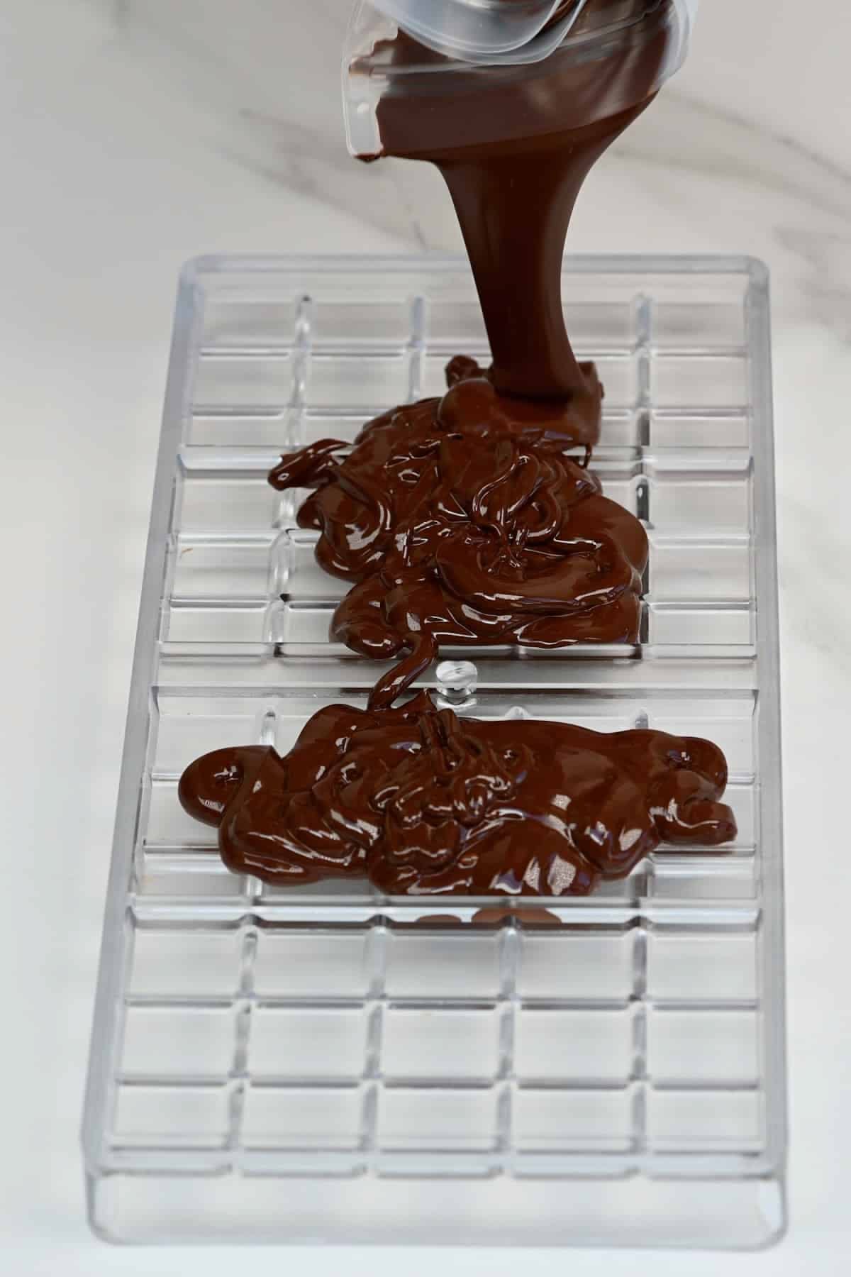 Melted dark chocolate being poured from a container into a clear plastic candy mold with grid compartments