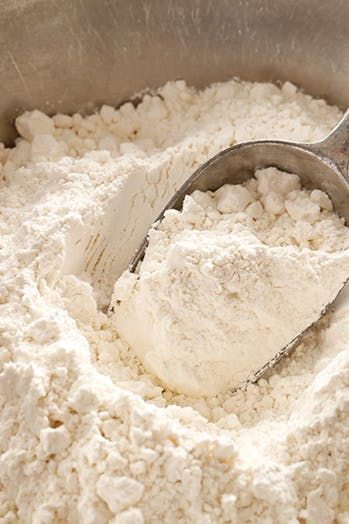 Close-up of white flour in a metal scoop, surrounded by a pile of flour.