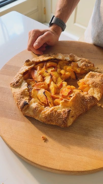 Baked peach galette on a wooden cutting board with a hand resting nearby.