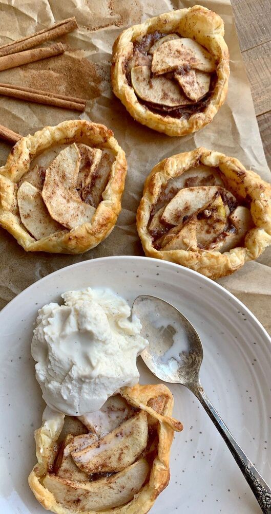 One individual apple galette on a white plate with a scoop of vanilla ice cream, next to two more galettes on brown…