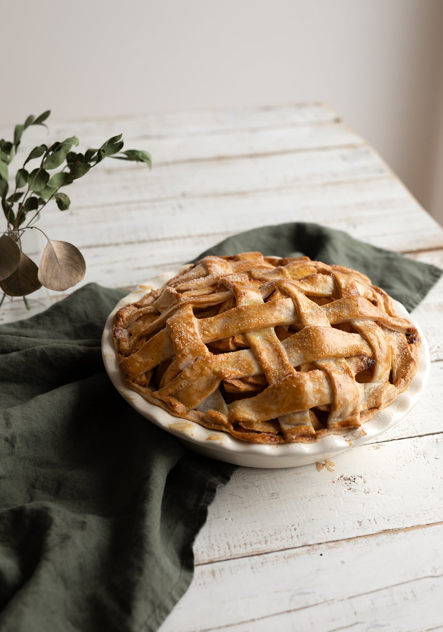 Baked apple pie with a lattice crust in a white pie dish, sitting on a dark green linen on a white wooden table