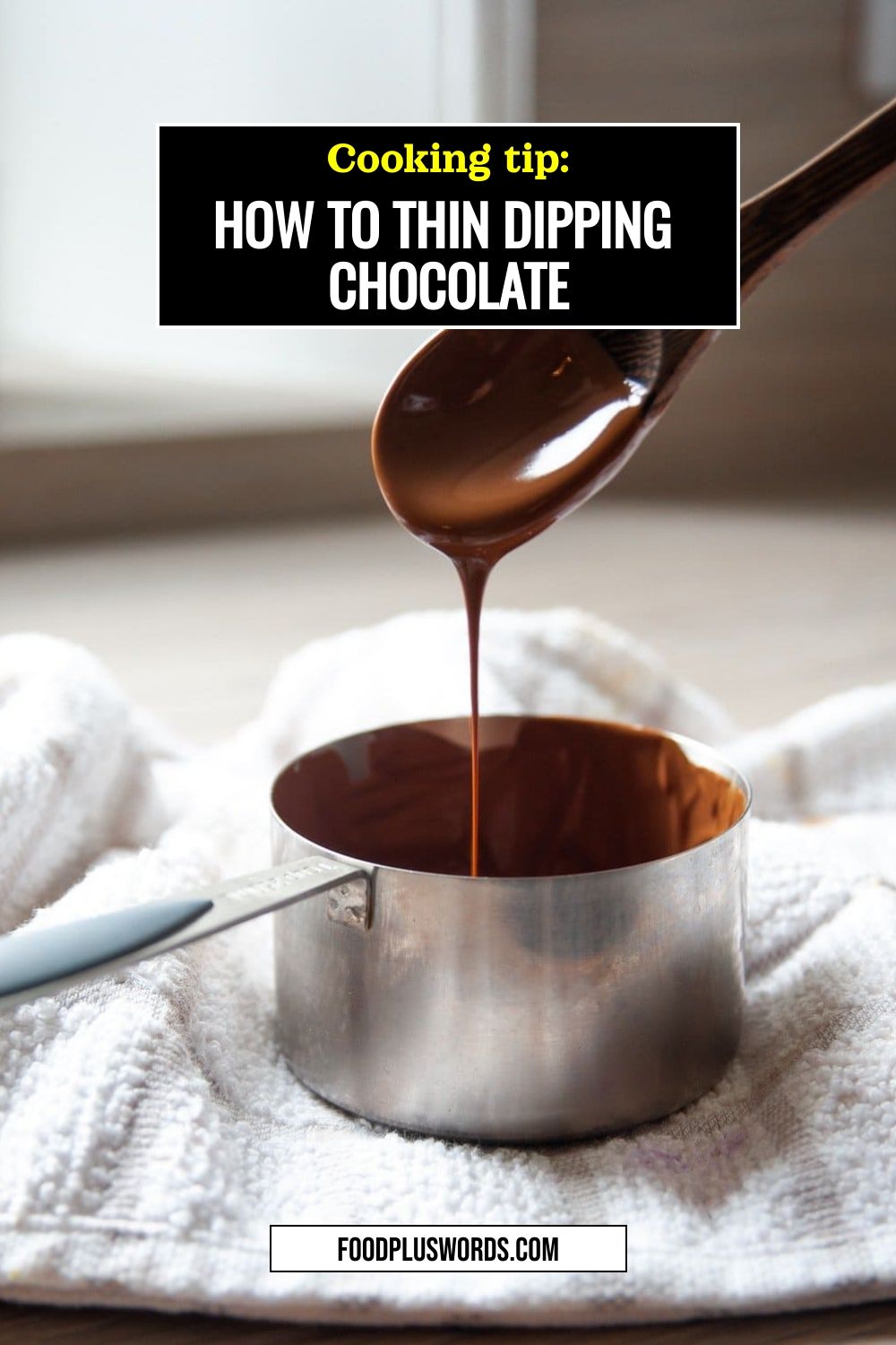 Melted chocolate pouring from a wooden spoon into a metal measuring cup, resting on a white towel.