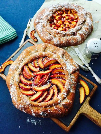 Two baked fruit galettes, one with sliced peaches arranged in a swirl, and another with diced fruit, on a dark blue surface.