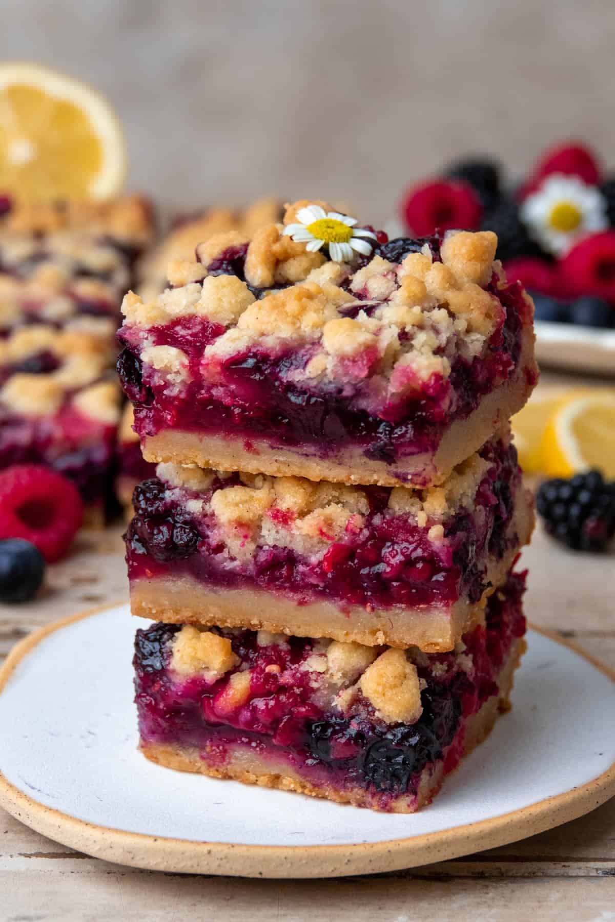 Three berry crumble bars stacked on a white plate, with a small white flower on top of the uppermost bar.