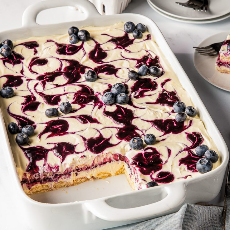 Blueberry tiramisu in a white rectangular baking dish, with one square piece removed. A swirl of blueberry sauce is on top.
