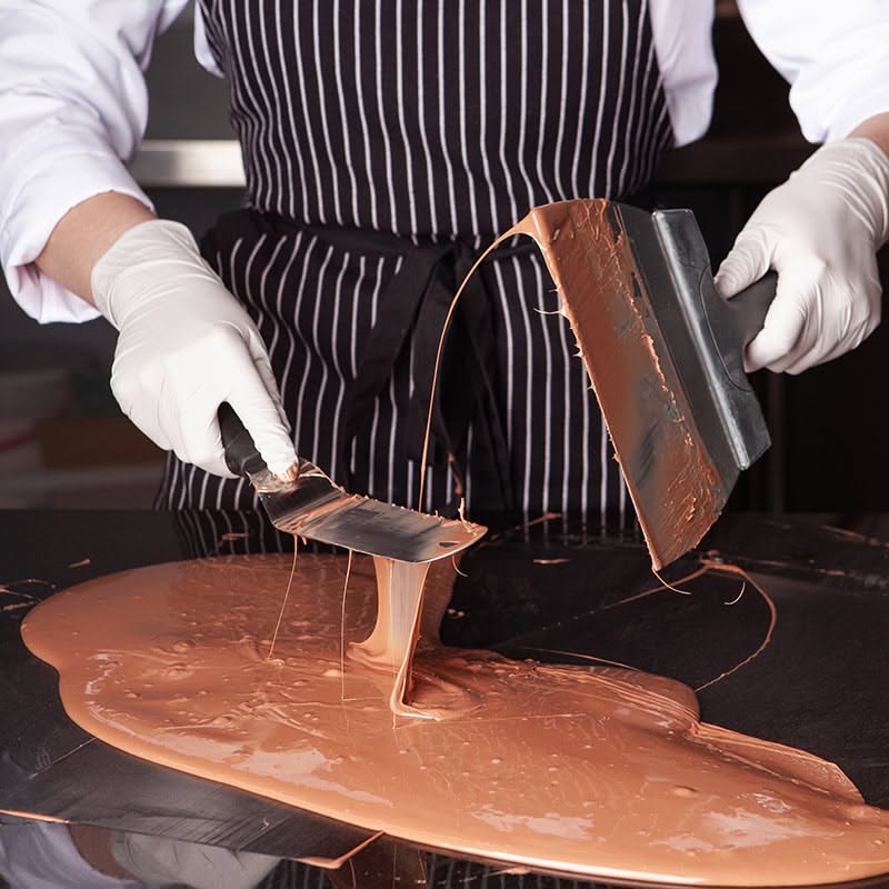 Gloved hands of a person in a striped apron spreading melted chocolate on a dark surface with two spatulas.