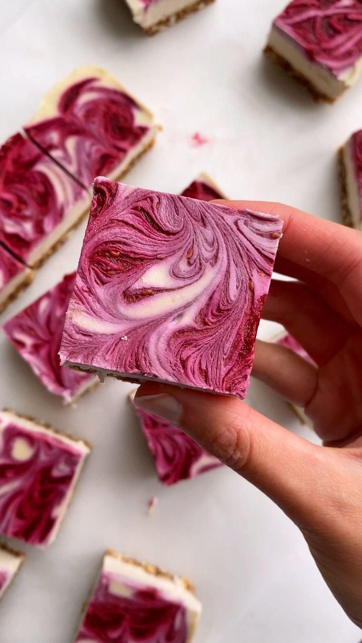 Hand holding a square vegan raspberry cheesecake bar with swirled pink and white topping, other bars on a white surface.
