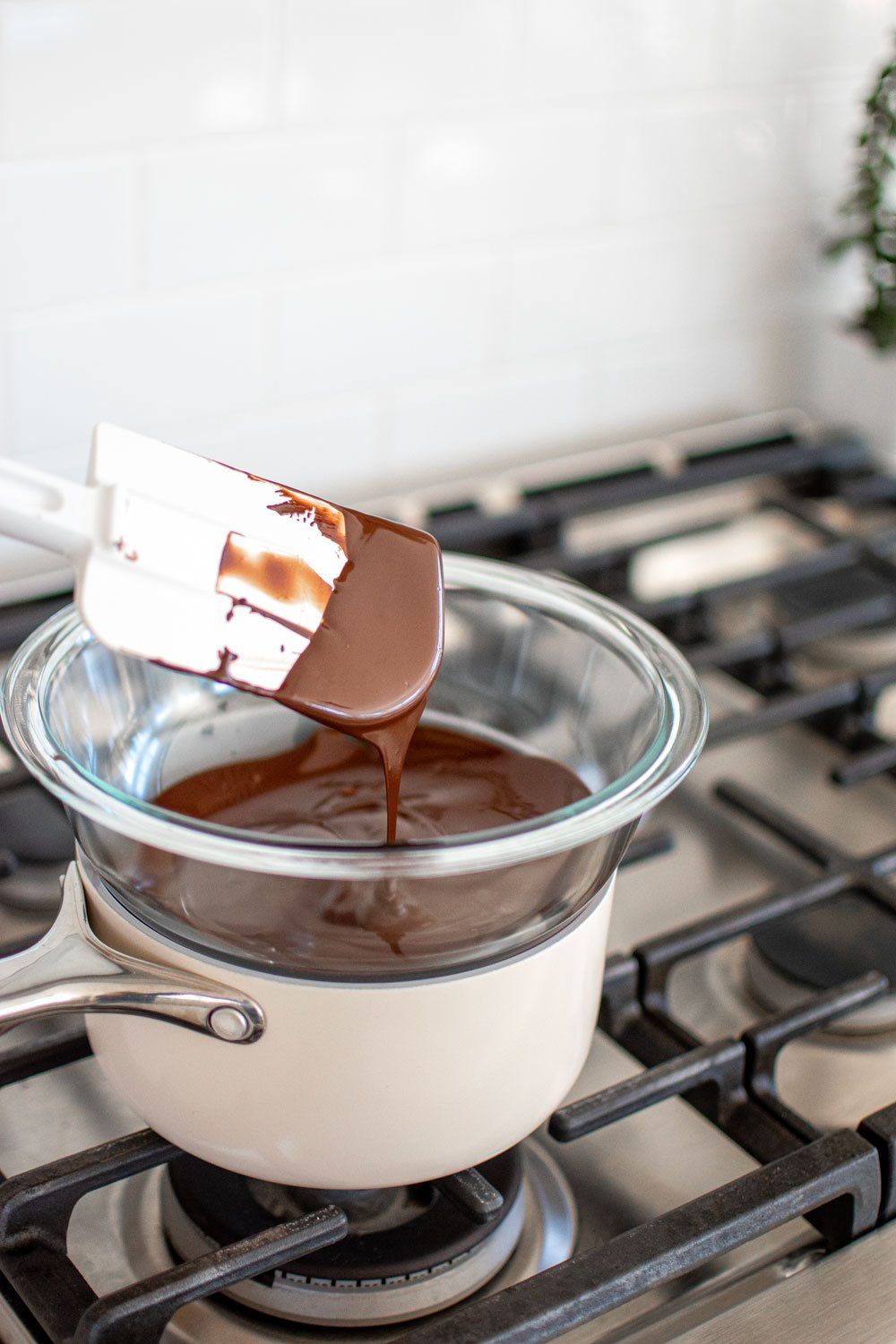 Melted chocolate in a glass bowl over a saucepan on a stovetop, with a white spatula dipping into it