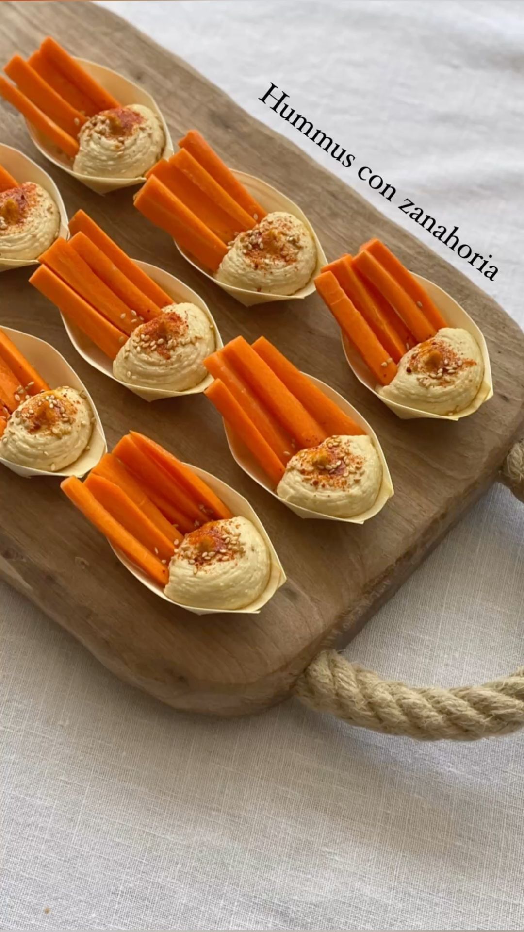 Several small boat-shaped dishes containing hummus and carrot sticks, arranged on a wooden board.
