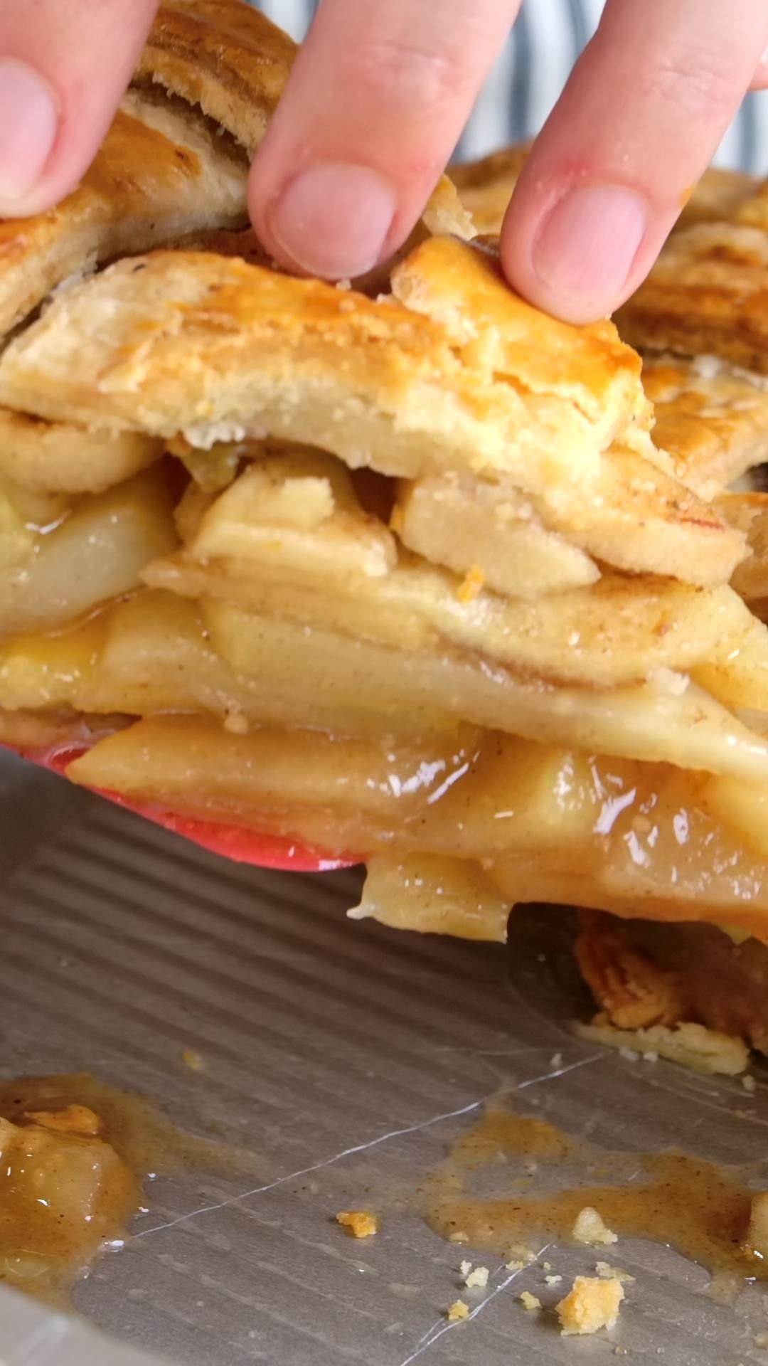 Close-up of a person's fingers lifting a piece of apple pie, revealing thick layers of spiced apple filling.