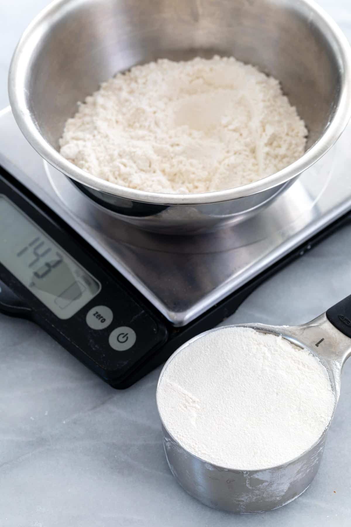 Bowl of flour on a digital kitchen scale showing 143 grams, with a measuring cup of flour nearby