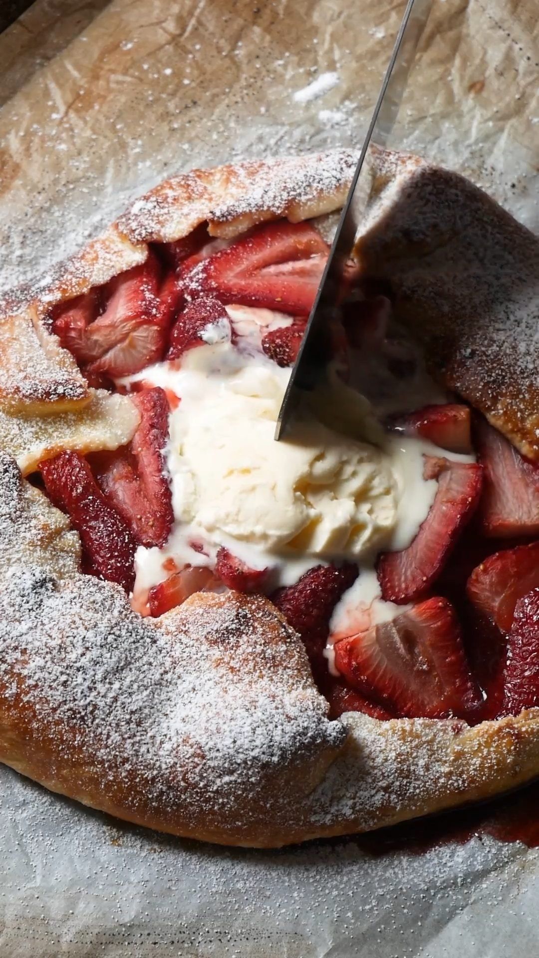 A knife cuts into a warm strawberry galette topped with melting vanilla ice cream and powdered sugar.