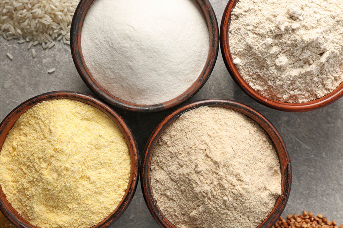 Four bowls of different types of flour, with whole grains like rice and buckwheat scattered nearby