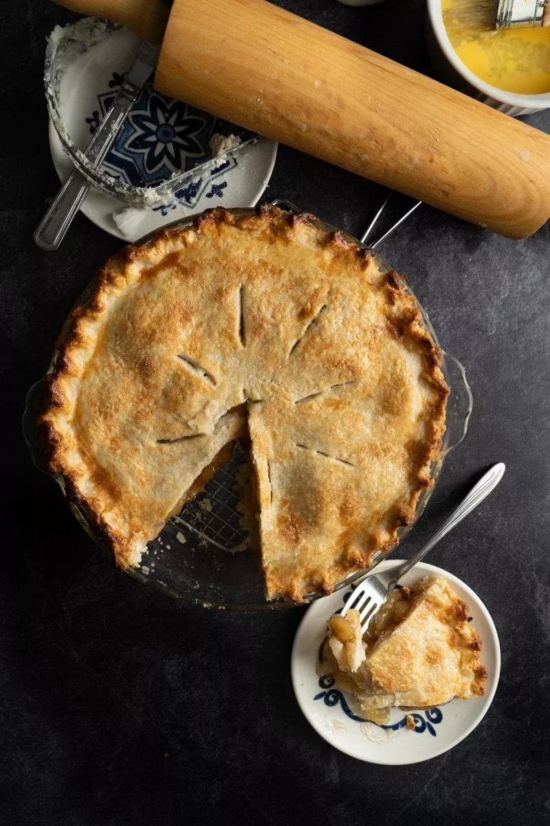 Baked apple pie in a glass dish with one slice on a small plate. A rolling pin and pastry blender are nearby.