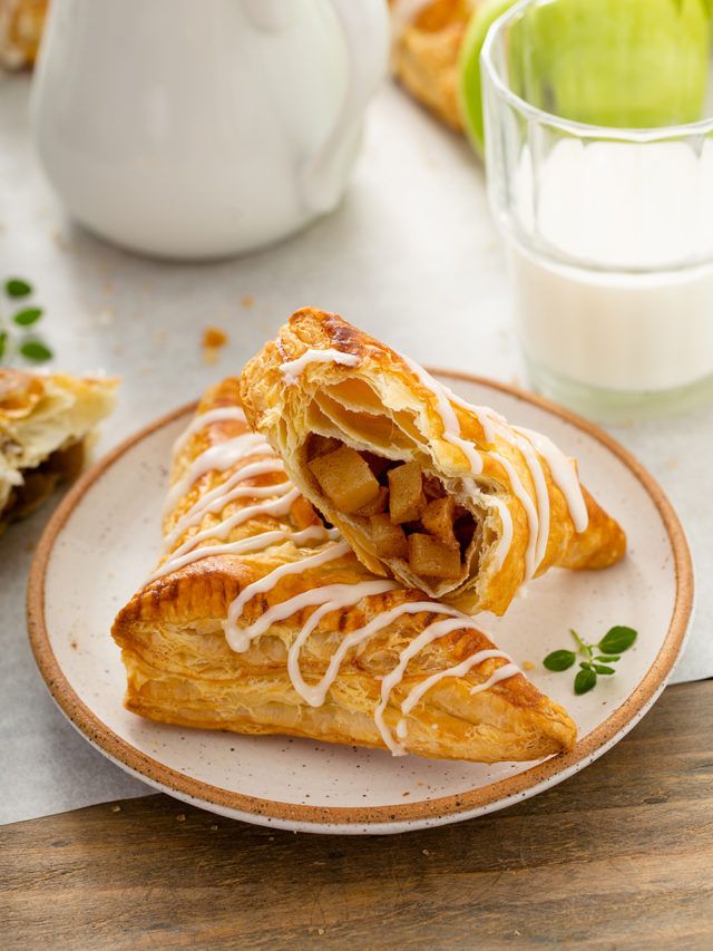 Two apple turnovers on a speckled plate, one cut in half showing apple filling, with a white glaze drizzle.