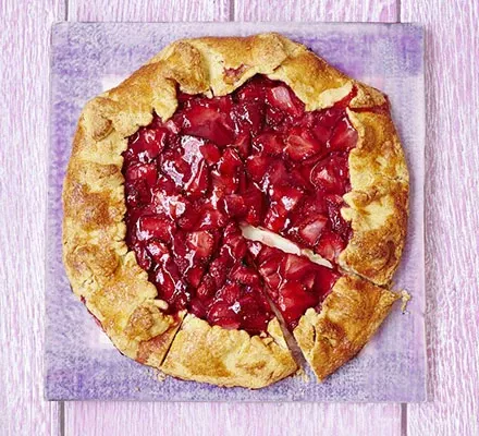 A whole strawberry galette, with a slice removed, on a light purple surface.
