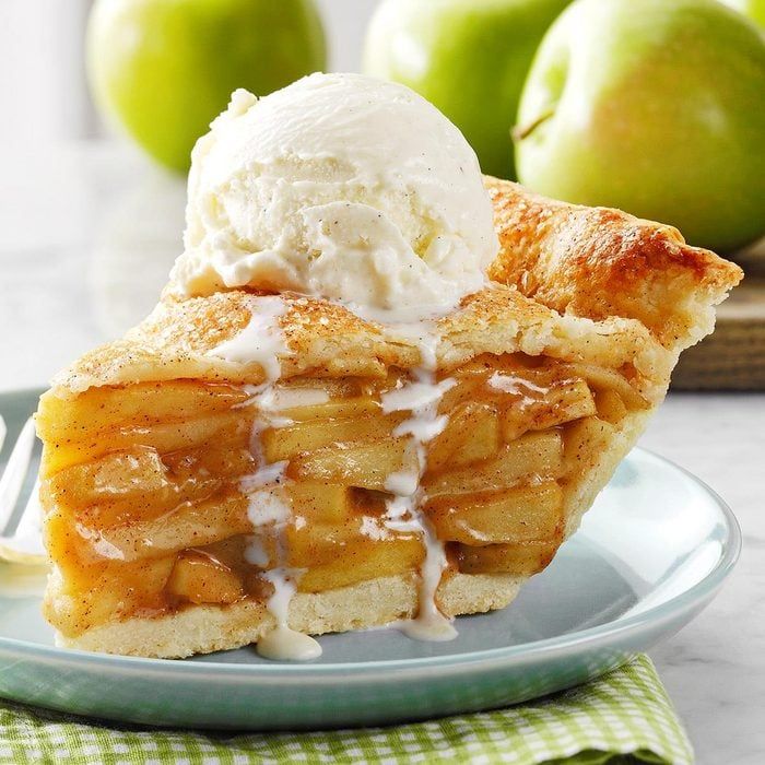 Slice of apple pie topped with melting vanilla ice cream on a light blue plate with green apples in background.