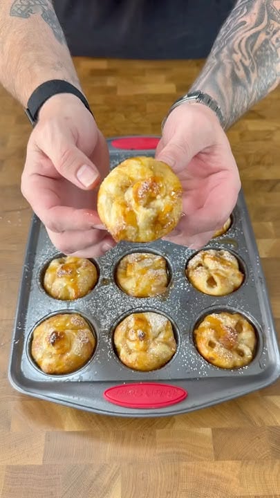 A person holds up a small, golden-baked apple galette, covered in glaze, above a muffin tin with several more.