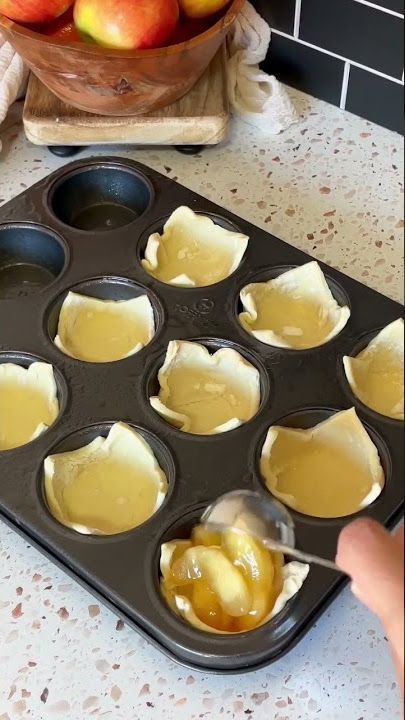 A hand uses a spoon to add cooked apple filling into a pastry-lined muffin tin cavity.