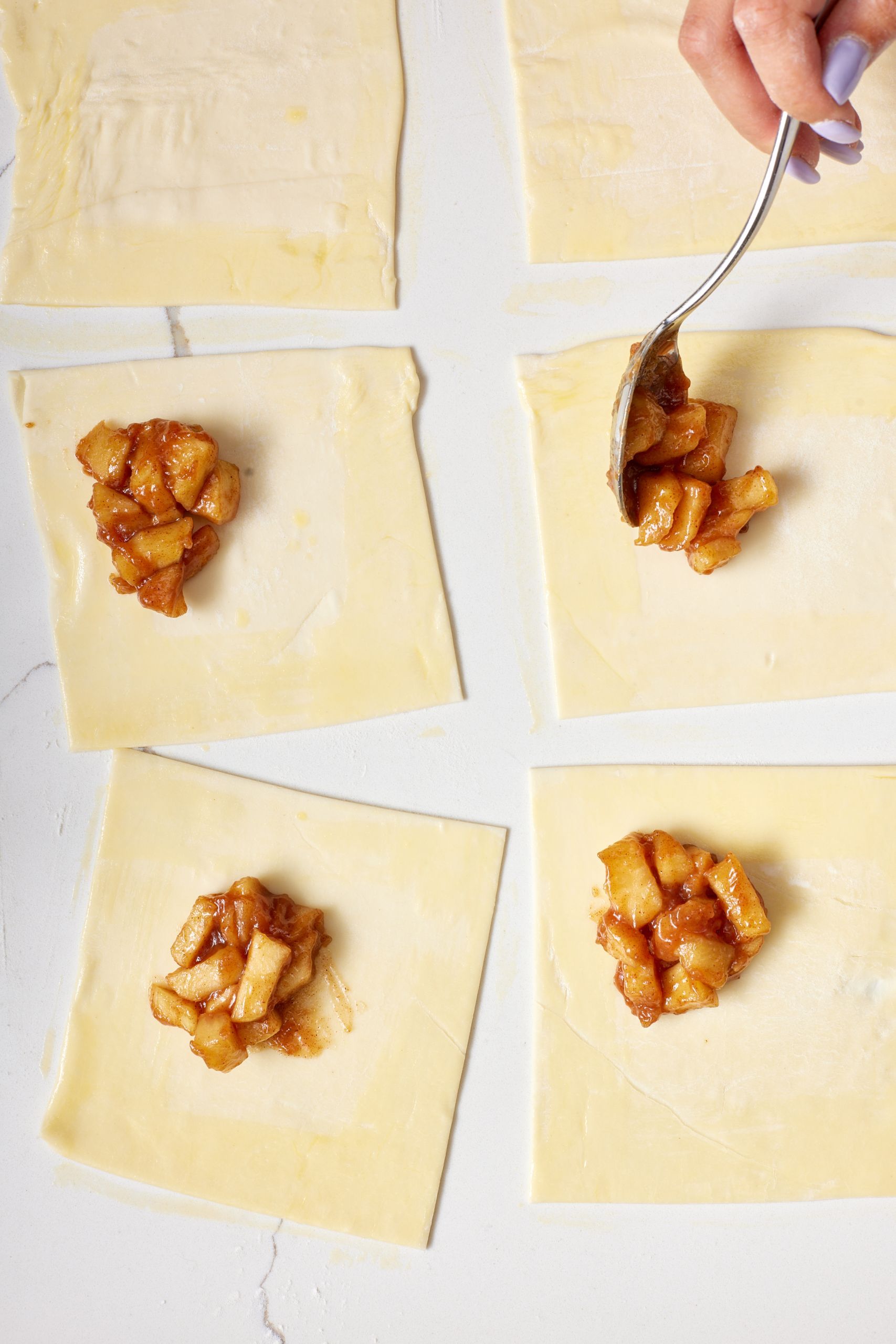 Overhead view of a hand scooping cooked apple filling onto squares of pastry dough on a white surface.