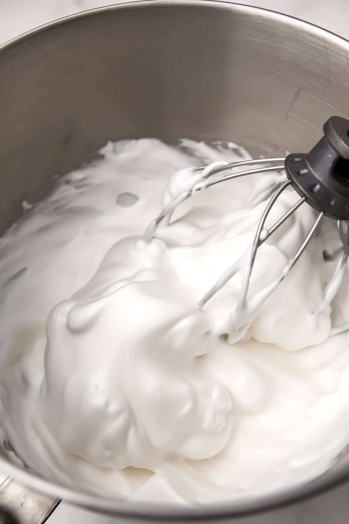 Close-up of thick, white, whipped aquafaba in a metal stand mixer bowl with a whisk attachment.