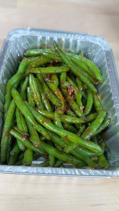 Close-up of a foil pan filled with baked green beans coated in a reddish-brown seasoning.