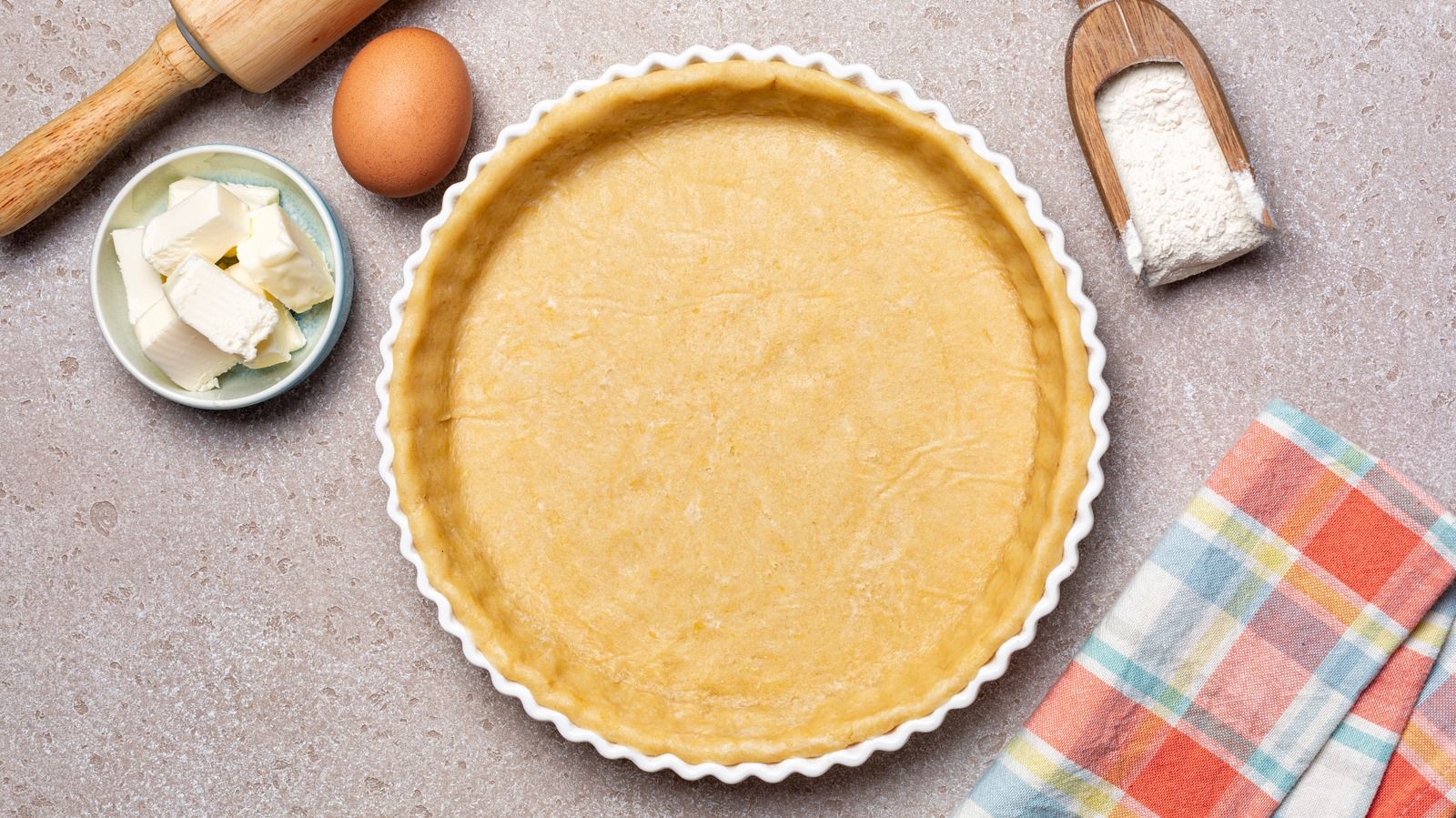 Unbaked tart crust in a white fluted tart pan, with ingredients like butter, an egg, and flour beside it.