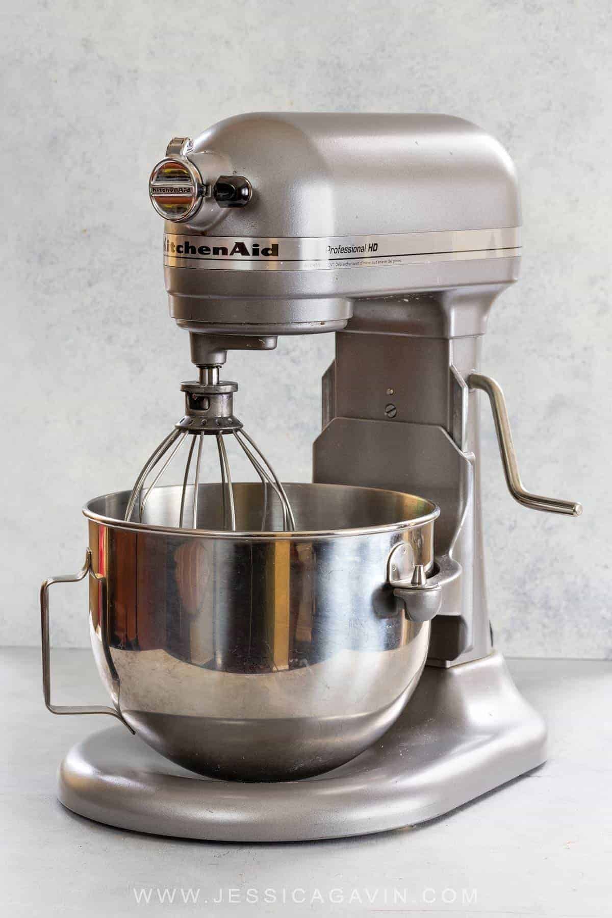 Silver stand mixer with a stainless steel bowl and whisk attachment on a light grey surface